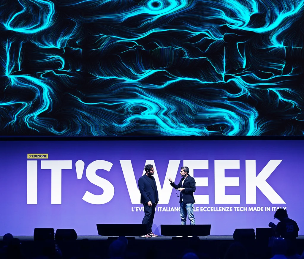 Due uomini in piedi sul palco di un evento tech, uno con microfono, davanti a un grande maxischermo che mostra un'opera dalle linee e dai vortici azzurri su fondo nero.