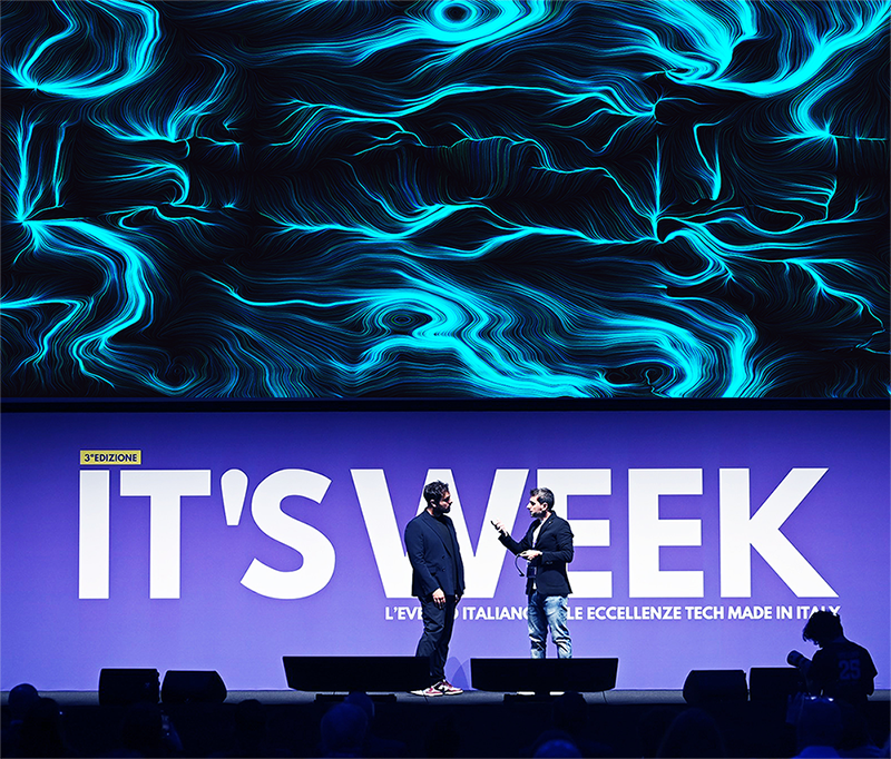 Due uomini in piedi sul palco di un evento tech, uno con microfono, davanti a un grande maxischermo che mostra un'opera dalle linee e dai vortici azzurri su fondo nero.