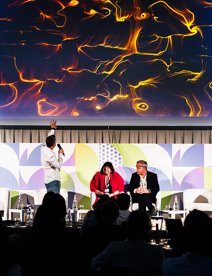 Relatore sul palco con opera BrainArt® arancione sullo schermo e panel di ospiti seduti durante una convention aziendale