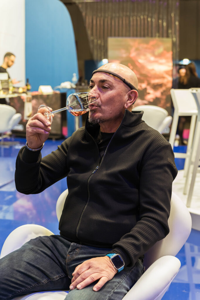 Un uomo seduto in poltrona, con un sensore EEG sulla fronte, porta alle labbra un calice di vino rosato; alle spalle lo stand di una fiera dai toni blu.
