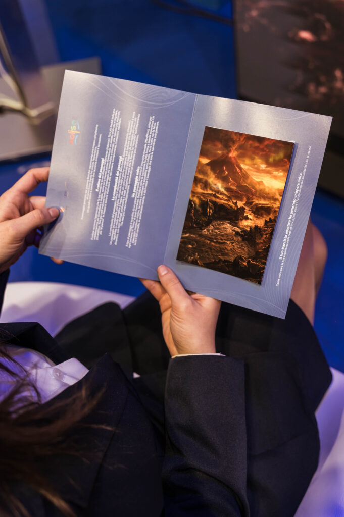 Le mani di una persona seduta tengono aperto un pieghevole BrainArt® con un testo sulla pagina sinistra e, sulla destra, la stampa di un paesaggio dai toni infuocati con un vulcano.