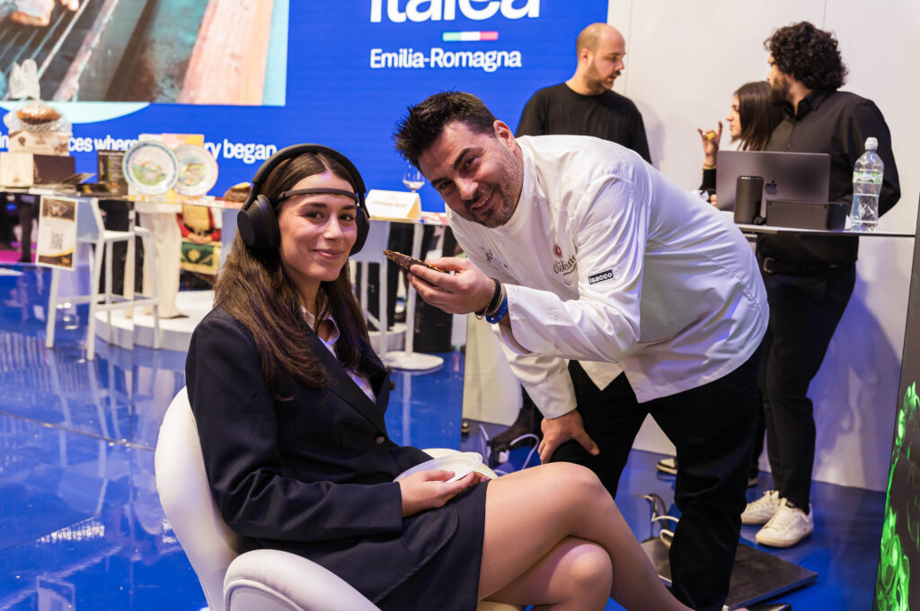 Una giovane donna seduta, con cuffie e un sensore EEG sulla fronte, tiene un piattino in grembo mentre uno chef in giacca bianca le porge un piccolo assaggio; sullo sfondo lo stand di una fiera.