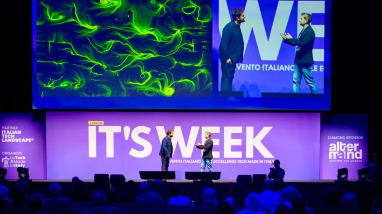 Il palco di una convention con la scritta "IT'S WEEK" e "3ª edizione" su fondo viola. Due uomini in piedi al centro del palco dialogano. Sopra di loro un grande schermo mostra, a sinistra, un'opera dalle fitte linee e dai vortici verdi su fondo scuro e, a destra, la ripresa live dei due relatori accanto alla scritta "WE". Ai lati del palco i loghi "Italian Tech Landscape", "LaTech #MadeinItaly" e "AlterMind". In basso, in controluce, il pubblico in platea.