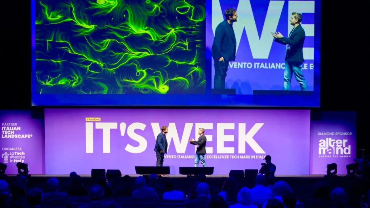 Il palco di una convention con la scritta "IT'S WEEK" e "3ª edizione" su fondo viola. Due uomini in piedi al centro del palco dialogano. Sopra di loro un grande schermo mostra, a sinistra, un'opera dalle fitte linee e dai vortici verdi su fondo scuro e, a destra, la ripresa live dei due relatori accanto alla scritta "WE". Ai lati del palco i loghi "Italian Tech Landscape", "LaTech #MadeinItaly" e "AlterMind". In basso, in controluce, il pubblico in platea.