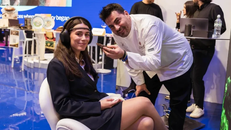 Una giovane donna in tailleur scuro è seduta su una poltroncina bianca con grandi cuffie sulle orecchie e un sensore appoggiato sulla fronte, e tiene in grembo un piattino; accanto a lei un uomo in giacca da cuoco bianca, sorridente e chino verso l'obiettivo, le porge un piccolo assaggio. Sullo sfondo un pannello blu con la scritta "Italea Emilia-Romagna" e altre persone attorno a un banco con un computer.