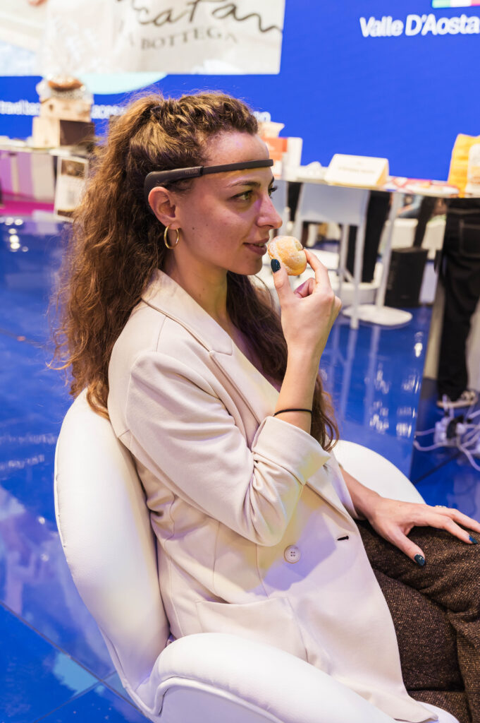 Una donna seduta in poltrona, con un dispositivo EEG sulla fronte e un abito color crema, avvicina al viso un piccolo dolce; alle spalle lo stand di una fiera dai toni blu.