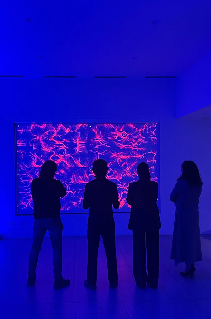 In una sala illuminata di blu intenso, quattro persone in piedi e in controluce osservano un grande schermo a parete. Sullo schermo un'opera dalle fitte linee e dai vortici rosa e magenta su fondo scuro. Le sagome delle persone si stagliano scure davanti alla luce; il pavimento chiaro riflette i toni blu dell'ambiente.
