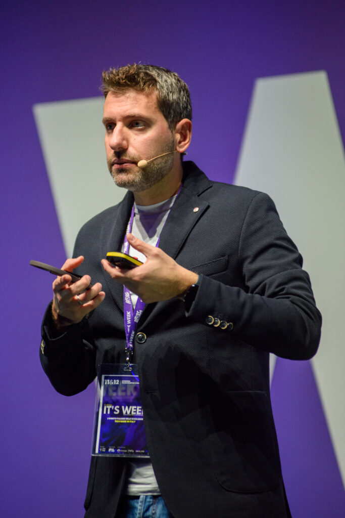 Un uomo parla sul palco di una conferenza, ripreso a mezza figura, con un microfono ad archetto e un'espressione concentrata. Indossa una giacca scura su maglietta chiara e tiene in mano due piccoli telecomandi. Al collo un pettorale con la scritta "IT'S WEEK" e le date "11 & 12". Alle sue spalle un fondale viola con una grande lettera bianca.