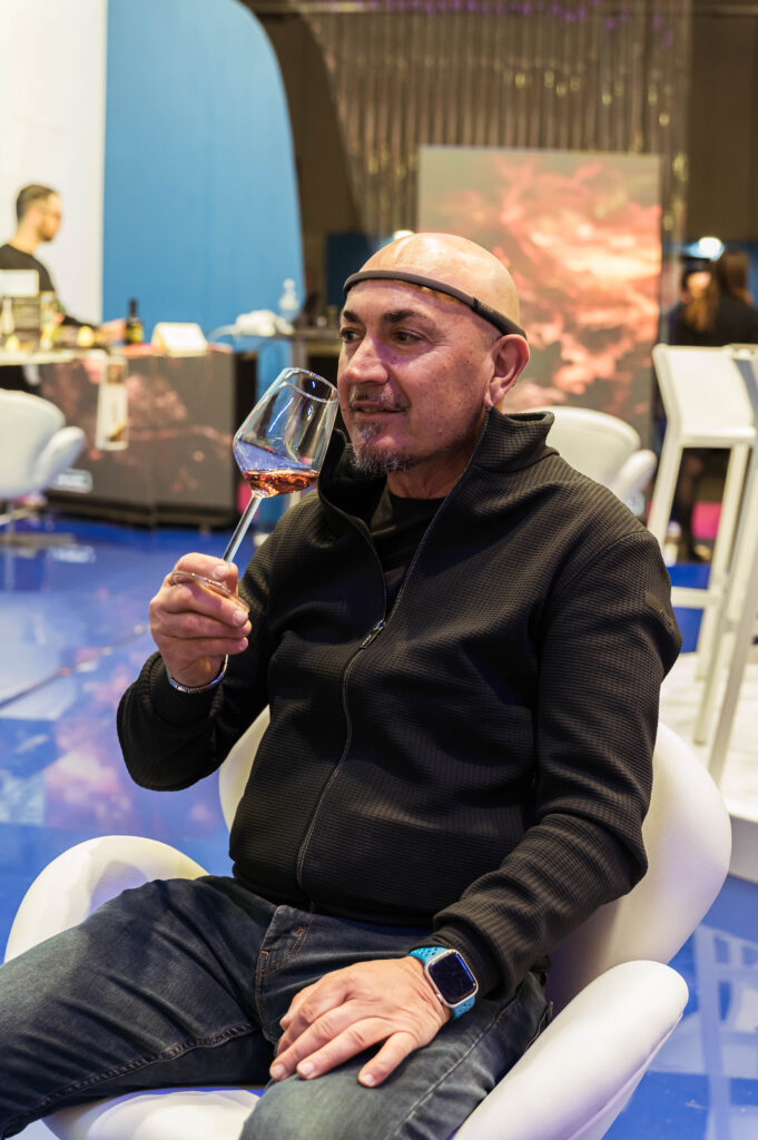 Un uomo con il capo rasato e un sensore EEG sulla fronte, seduto in una poltroncina bianca, tiene in mano un calice con un vino di colore rosato e guarda di lato. Indossa una felpa nera con zip e jeans. Alle sue spalle, sfocati, un pavimento lucido blu, sedute bianche, un bancone e uno schermo con un'opera.