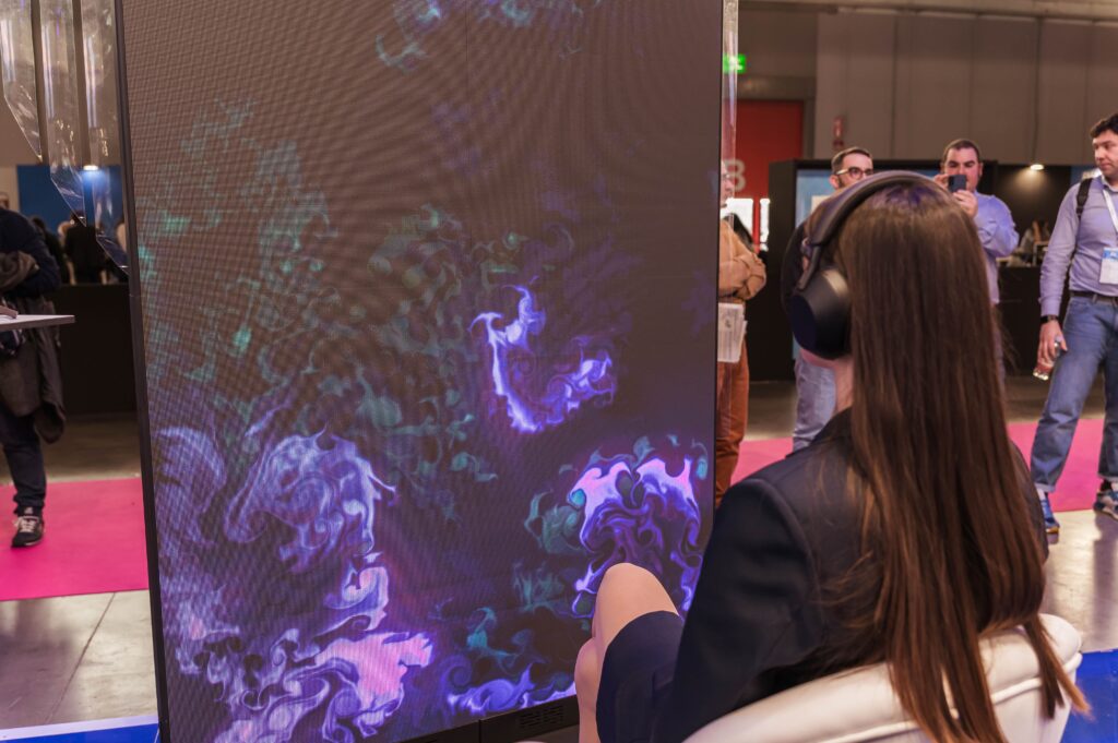 Un grande schermo mostra un'opera dalle fitte linee e dai vortici viola, rosa e azzurri su fondo scuro; in primo piano, di spalle, una persona con le cuffie osserva lo schermo, mentre sullo sfondo si intravedono altri visitatori in un ambiente espositivo.
