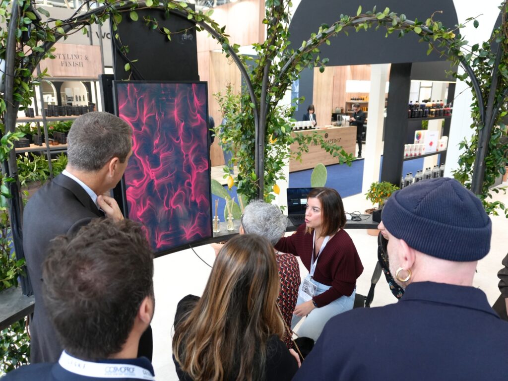 In uno stand fieristico a tema beauty, decorato con archi di verde e scaffali di prodotti, una hostess con badge seduta parla a un gruppo di visitatori raccolti intorno a lei. Accanto, uno schermo verticale mostra un'opera dalle linee e dai vortici rosa e magenta su fondo scuro. Sulla sinistra un'insegna "Styling & Finish"; sullo sfondo un bancone in legno e diffusori a bastoncini. Uno dei presenti porta un pettorale con la scritta "Cosmoprof".