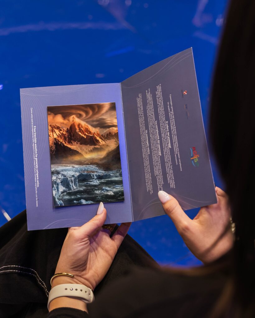 Le mani di una donna tengono aperto un pieghevole dai toni viola-blu. Sulla pagina di sinistra è inserita un'immagine figurativa che raffigura una montagna innevata illuminata dal sole sopra una cascata e nubi mosse; sulla pagina di destra un testo stampato, alcuni piccoli simboli e un logo colorato in basso. Lo sfondo è un pavimento blu.