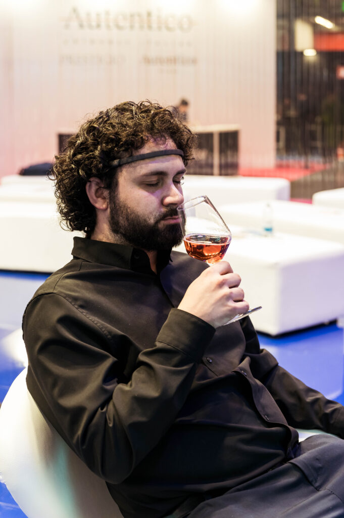 Un uomo con capelli ricci scuri e barba, seduto e ripreso di profilo, indossa un sensore EEG sulla fronte e porta al naso un calice con un vino di colore rosato, con gli occhi abbassati. Indossa una camicia nera. Alle sue spalle, sfocati, sedute bianche, un pavimento lucido blu e un'insegna con la scritta "Autentico".