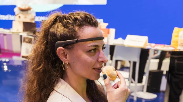 Una donna con lunghi capelli ricci castani, ripresa di profilo, indossa sulla fronte una fascia scura con sensore (sul lato la scritta «muse») e avvicina al viso un piccolo dolce spolverato di zucchero a velo che tiene tra le dita. Indossa una giacca chiara e orecchini a cerchio. Sullo sfondo, sfocati, uno stand fieristico con una parete blu, ripiani con prodotti confezionati e sgabelli.