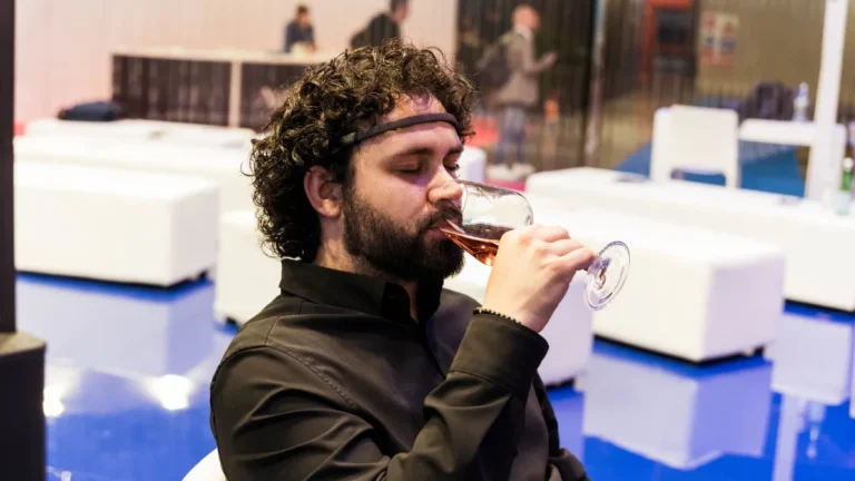 Un uomo con capelli ricci scuri e barba, seduto e ripreso di profilo, indossa un sensore EEG sulla fronte e, con gli occhi chiusi, beve da un calice un vino di colore rosato. Indossa una camicia nera. Alle sue spalle, sfocati, sedute e pouf bianchi, un pavimento lucido blu e alcune persone in piedi in un'area lounge.