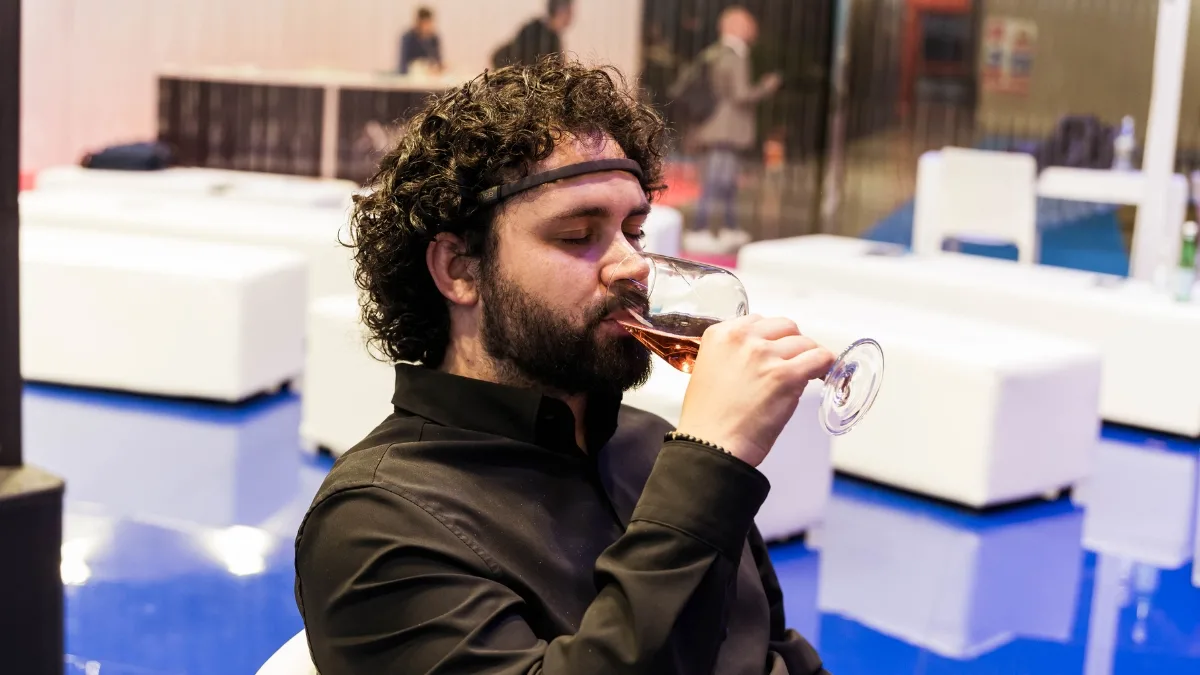 Un uomo con capelli ricci scuri e barba, seduto e ripreso di profilo, indossa un sensore EEG sulla fronte e, con gli occhi chiusi, beve da un calice un vino di colore rosato. Indossa una camicia nera. Alle sue spalle, sfocati, sedute e pouf bianchi, un pavimento lucido blu e alcune persone in piedi in un'area lounge.