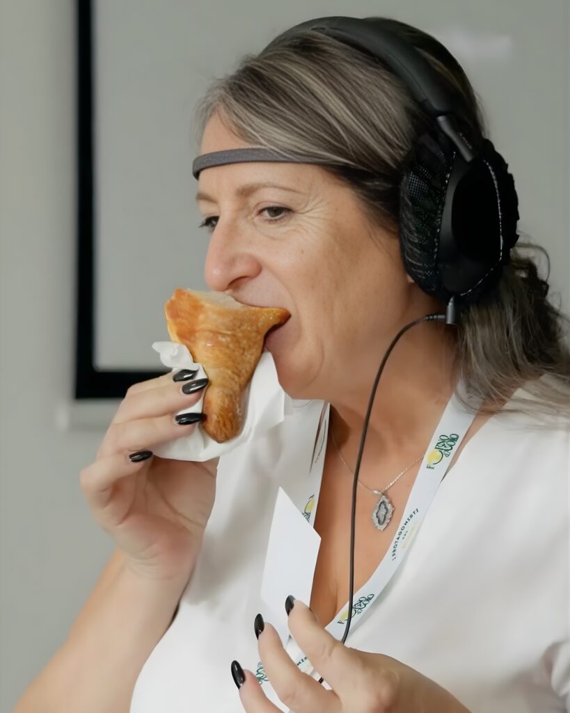 Una donna con il sensore appoggiato sulla fronte e grandi cuffie sulle orecchie addenta un cornetto avvolto in un tovagliolo, tenendolo con entrambe le mani; indossa una camicia bianca con un badge al collo e, sullo sfondo, una parete chiara.