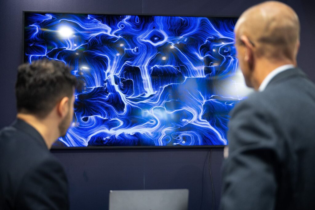 Due uomini in giacca scura, ripresi di spalle e di profilo, osservano da vicino uno schermo a parete. Sullo schermo un'opera dalle fitte linee e dai vortici blu e bianchi luminosi su fondo scuro, con alcuni punti di luce più intensi. La parete intorno è scura; sotto lo schermo si intravede lo schienale di una seduta.