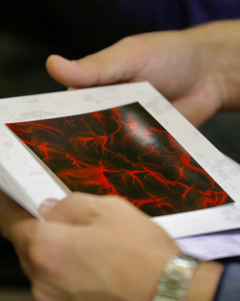 Due mani reggono una cartella chiara aperta al cui interno è applicata la stampa di un'opera dalle fitte linee rosse su fondo nero; una terza mano, sulla destra, sostiene il bordo della cartella. Lo sfondo è scuro e sfocato.