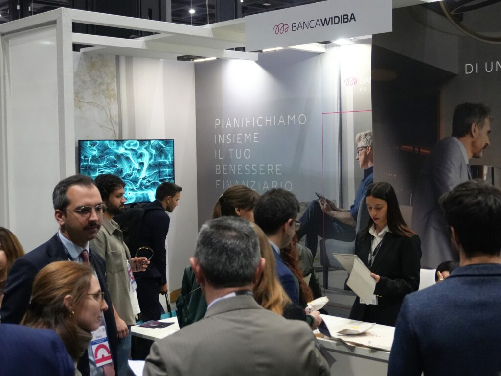Vista ampia di uno stand fieristico affollato con l'insegna "BANCA WIDIBA". Sulla sinistra, uno schermo mostra un'opera dalle linee e dai vortici turchesi su fondo scuro; al centro la scritta "Pianifichiamo insieme il tuo benessere finanziario" e un'immagine di persone. Numerosi visitatori in abito da lavoro conversano davanti a un bancone; una hostess con tablet parla con un ospite.