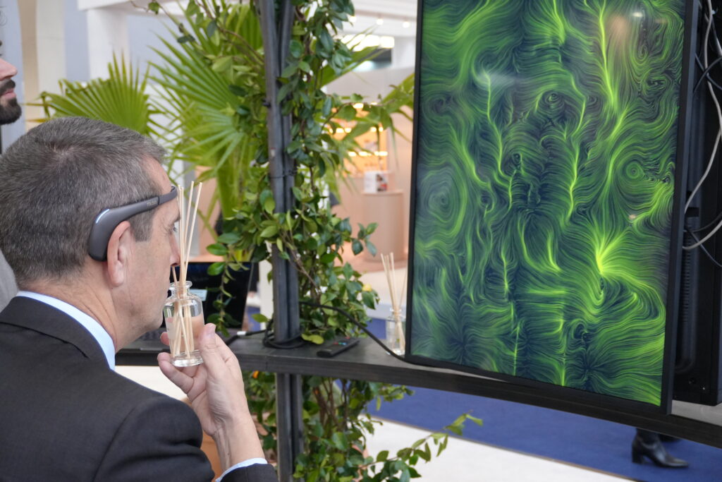 Un uomo di profilo, con il sensore appoggiato sulla fronte, tiene in mano una boccetta di vetro con bastoncini di legno e la avvicina al viso; davanti a lui un grande schermo verticale mostra un'opera dalle fitte linee e dai vortici verdi su fondo scuro, mentre attorno alla postazione ci sono piante verdi e, sullo sfondo, un padiglione fieristico.