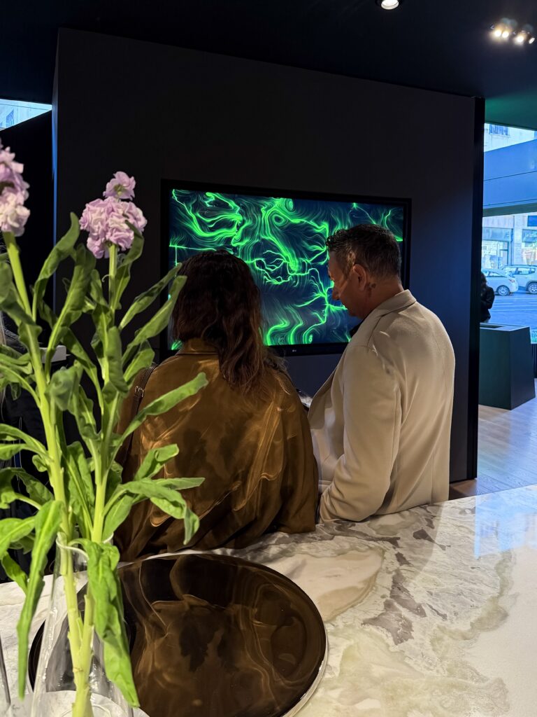 Due persone di spalle davanti a uno schermo che mostra un'opera dalle linee e dai vortici verdi su fondo scuro; in primo piano un piano in pietra chiara venata con un vaso di fiori lilla e una coppa metallica, sullo sfondo la vetrata dello showroom.