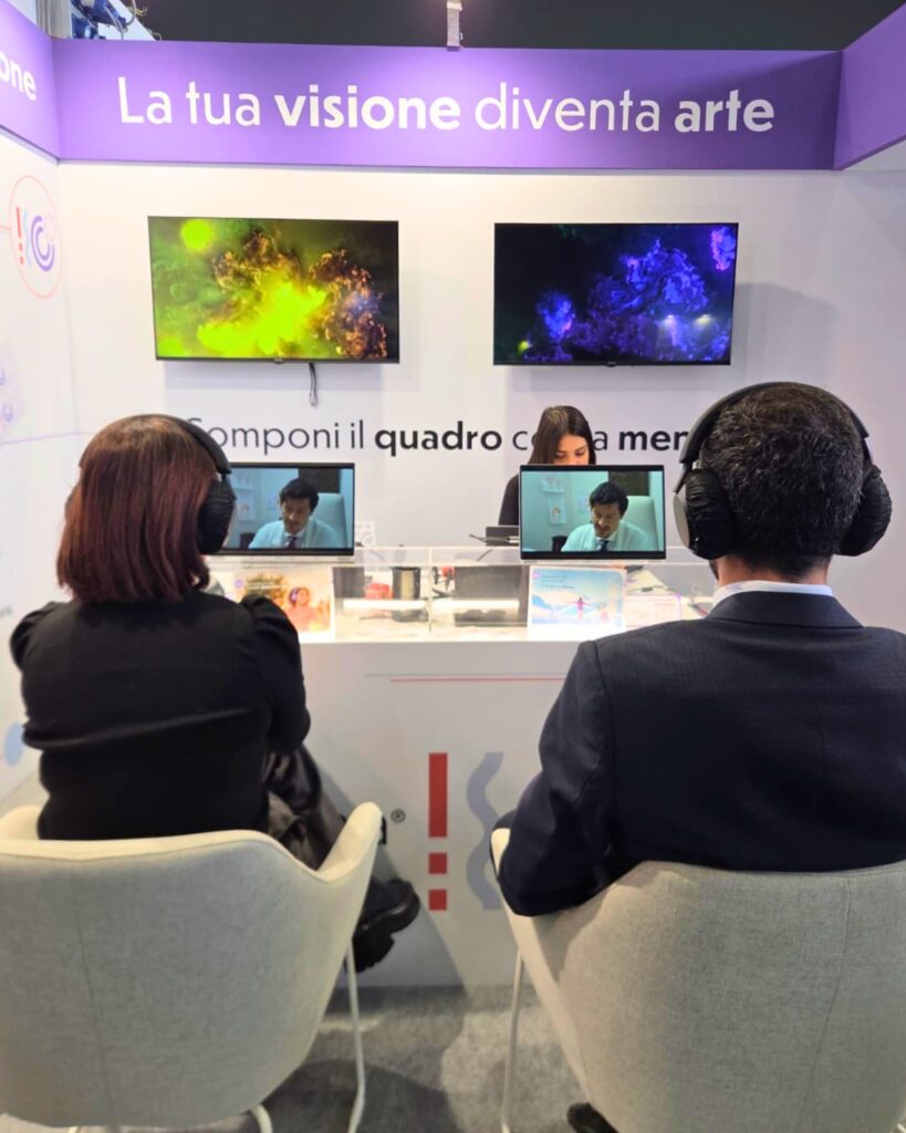 Due partecipanti seduti di spalle, con le cuffie, davanti alle postazioni dello stand Lo.Li. Pharma: sui monitor scorre un video, mentre gli schermi in alto mostrano due opere dalle linee e dai vortici, una verde e una viola; sopra il claim "La tua visione diventa arte".