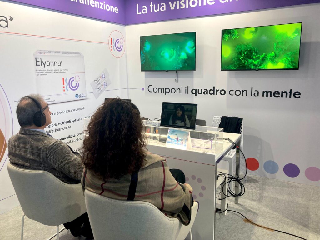Due partecipanti seduti di spalle, con le cuffie, guardano i monitor delle postazioni su cui scorre un video, allo stand Lo.Li. Pharma; sopra di loro due schermi mostrano opere dalle linee e dai vortici verdi e, accanto, il claim "Componi il quadro con la mente" e la parete dedicata a Elyanna.