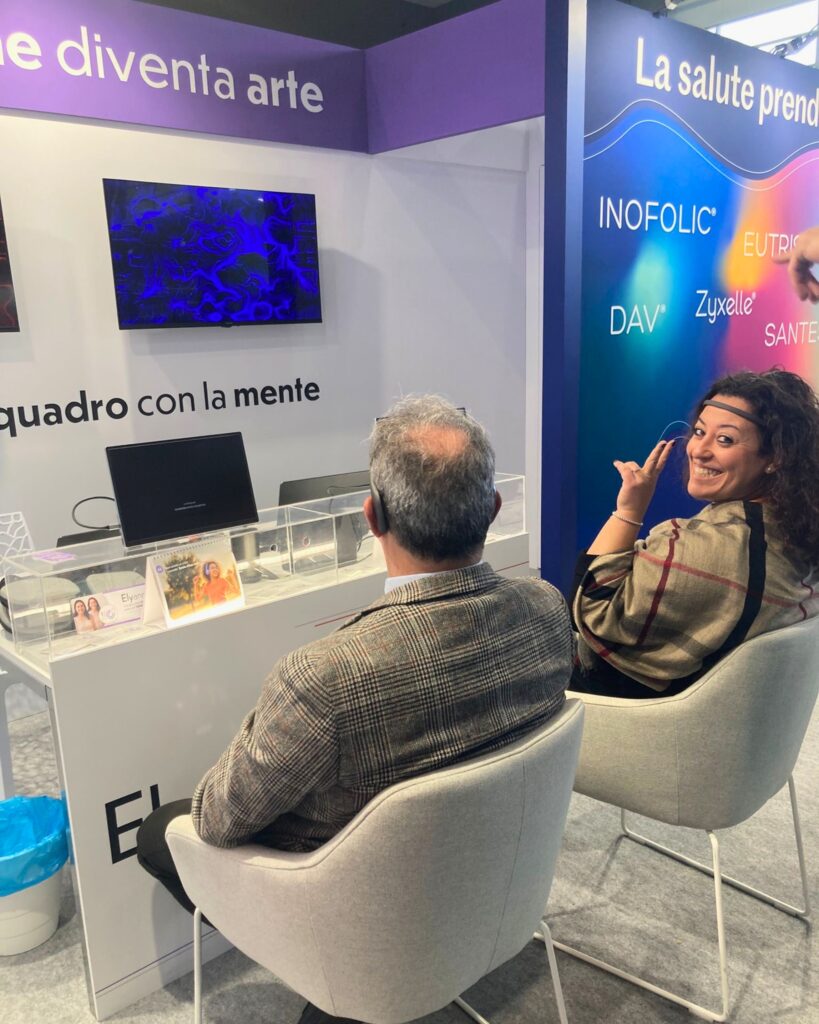 Una donna con un sensore EEG, seduta a una postazione dello stand Lo.Li. Pharma, si volta sorridendo verso l'obiettivo; accanto a lei un uomo di spalle guarda il monitor, mentre sullo schermo alle spalle è visibile un'opera dalle linee e dai vortici blu.