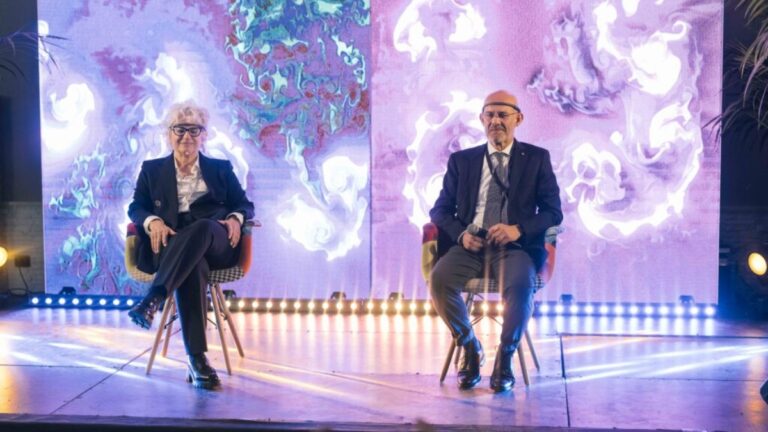 Due persone sedute su poltroncine sul palco di un evento aziendale, davanti a due grandi schermi che mostrano opere dalle linee e dai vortici bianchi, rosa e viola; ai loro piedi una fila di luci.