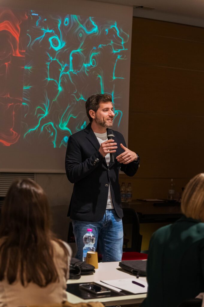 Un uomo in giacca scura, con microfono in mano, parla in piedi davanti alla platea seduta ai tavoli; alle sue spalle uno schermo mostra un'opera dalle linee e dai vortici turchesi e, sul lato sinistro, una porzione di un'altra opera rossa.