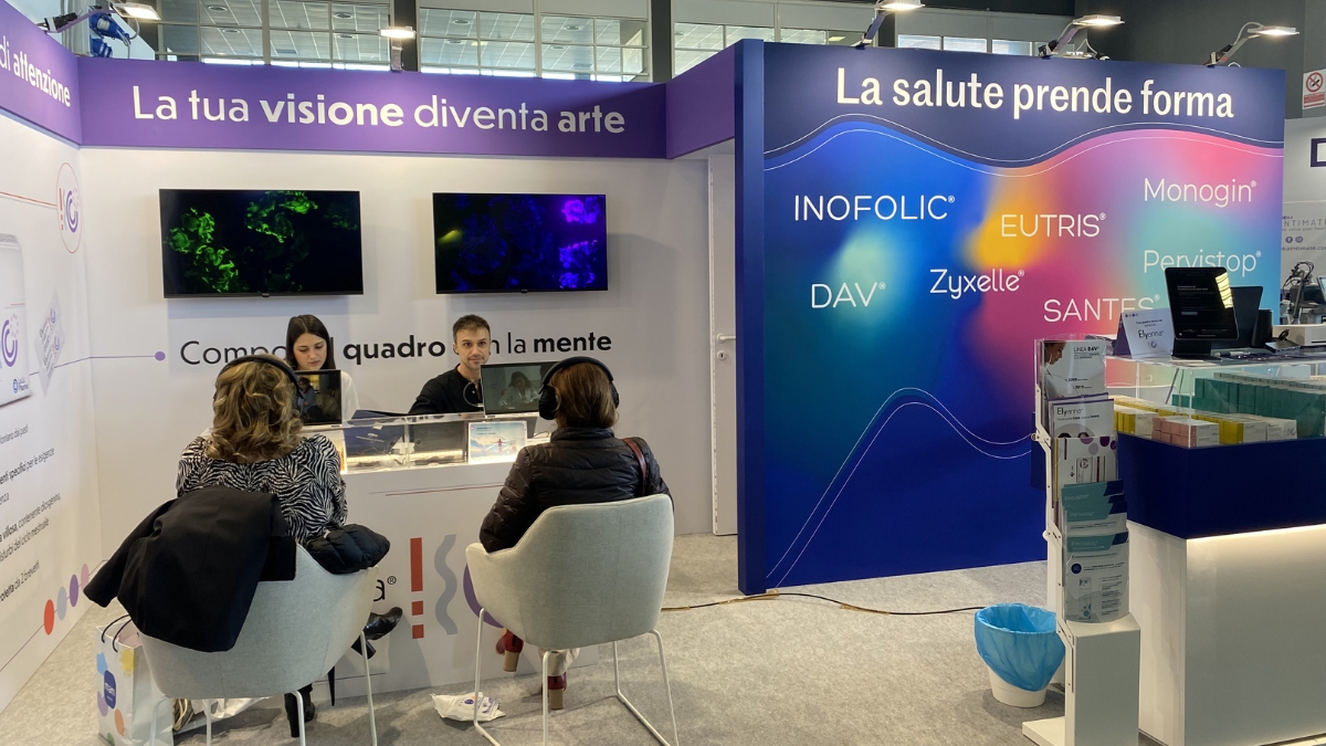 Lo stand di Lo.Li. Pharma al congresso SIGO, con i claim "La tua visione diventa arte" e "Componi il quadro con la mente", due schermi che mostrano opere dalle linee e dai vortici verdi e viola e due partecipanti sedute con le cuffie davanti alle postazioni; accanto la parete "La salute prende forma" con i marchi dell'azienda.