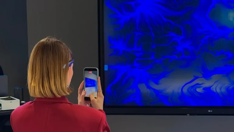 Una donna di spalle, in giacca bordeaux e con un sensore appoggiato sulla fronte, fotografa con lo smartphone un grande schermo a parete che mostra un'opera dalle fitte linee e dai vortici blu su fondo scuro; l'ambiente ha pareti grigie e, in basso, un piano in marmo chiaro.