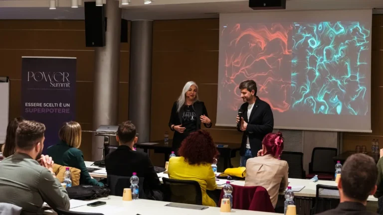 Due relatori in piedi parlano davanti a una platea seduta ai tavoli in una sala meeting; sullo schermo alle loro spalle sono proiettate due opere affiancate, una a linee rosse e una a linee azzurre su fondo scuro, mentre sulla sinistra un roll-up riporta la scritta "Power Summit — Essere scelti è un superpotere". In primo piano il pubblico di spalle con bottiglie d'acqua e quaderni sui tavoli.