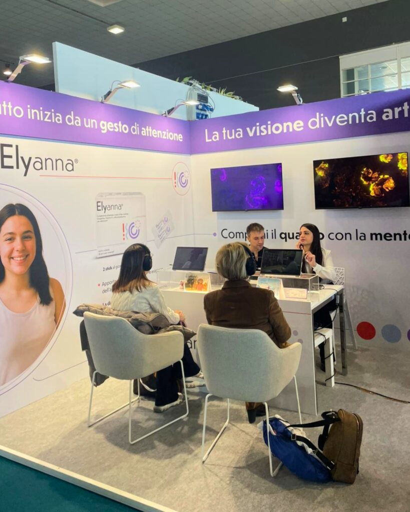 Lo stand di Lo.Li. Pharma con la parete dedicata a Elyanna e i claim "Tutto inizia da un gesto di attenzione" e "La tua visione diventa arte"; due partecipanti sedute con le cuffie davanti alle postazioni e, sugli schermi, due opere dalle linee e dai vortici, una viola e una dorata.
