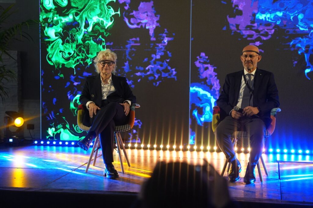 Su un palco, due relatori seduti su poltroncine distanziate: a sinistra una donna con giacca scura e occhiali, a destra un uomo con giacca scura, cravatta e copricapo chiaro. Alle loro spalle una grande proiezione a parete mostra due opere affiancate, una dalle volute e dai vortici verdi e una dai vortici blu e viola, su fondo scuro. Ai piedi del fondale una fila di luci gialle e blu; il pavimento riflette i fasci di luce. In basso, sfocata, la sagoma di una persona del pubblico.