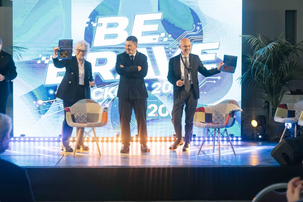 Tre persone sul palco davanti al maxischermo con la scritta "Be Brave" e il riferimento al Kick Off 2026: due di loro mostrano al pubblico i pieghevoli con le stampe delle proprie opere, una dorata e una rossa.