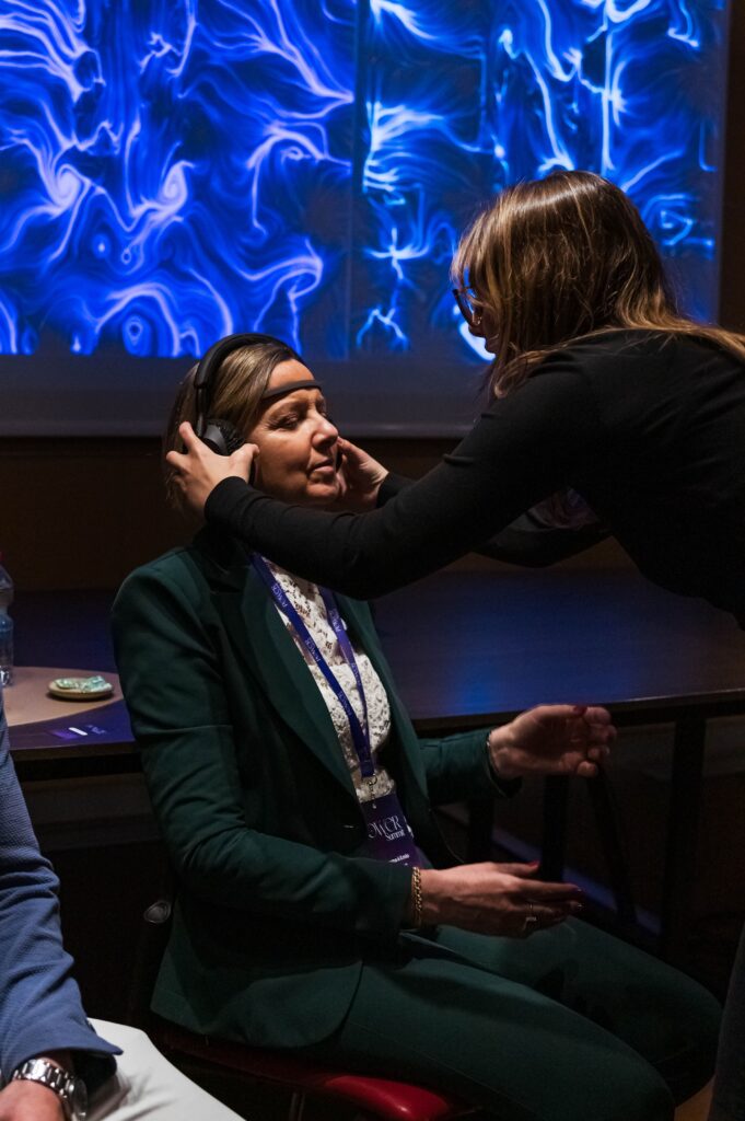 Una donna in piedi sistema con entrambe le mani le cuffie e il sensore EEG sulla testa di un'altra donna seduta, in giacca verde e con il pass al collo; alle loro spalle uno schermo mostra un'opera dalle linee e dai vortici blu.