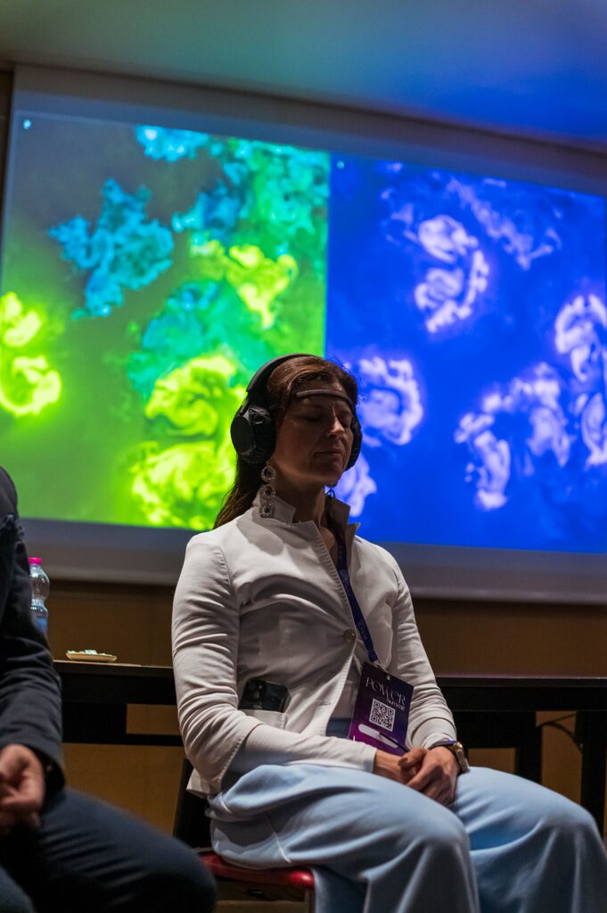 Una donna seduta a occhi chiusi, con un sensore EEG sulla fronte, le cuffie in testa, il pass del Power Summit al collo e le mani in grembo; alle sue spalle uno schermo con due opere affiancate, una dai vortici verdi e azzurri e una dai vortici blu e bianchi.