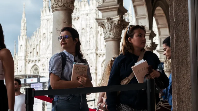 Alcune persone in coda sotto i portici di una piazza, delimitate da una transenna a nastro rosso; una donna con occhiali e camicia a righe tiene tra le braccia un libro con in copertina il nome dell'autore e il titolo, mentre accanto a lei altre persone attendono con dei pieghevoli chiari in mano; sullo sfondo, oltre le colonne, si vedono le guglie bianche del Duomo di Milano sotto un cielo nuvoloso.