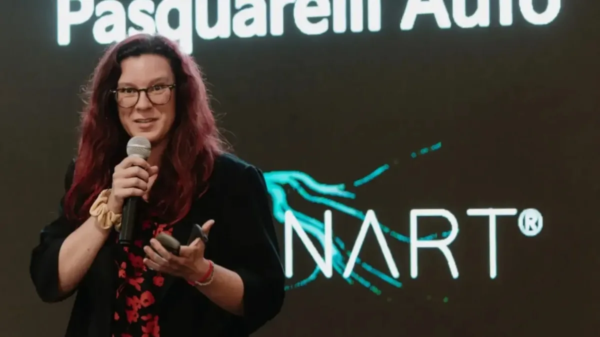 Una donna dai capelli rossi, con occhiali e abito scuro a fiori, parla al microfono tenendo in mano un telecomando; alle sue spalle uno schermo scuro mostra la scritta "Pasquarelli Auto" e il logo BrainArt® con la sua grafica a linee turchesi.