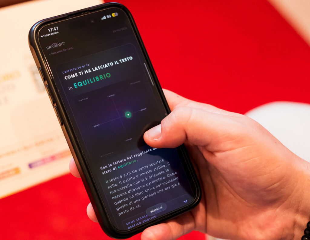 Una mano regge uno smartphone che mostra la schermata "L'effetto su di te – Come ti ha lasciato il testo: in EQUILIBRIO", con un grafico a due assi su fondo scuro e un punto verde luminoso posizionato al centro; sotto, un testo spiega che con la lettura si è raggiunto uno stato di equilibrio. Sullo sfondo un piano rosso.