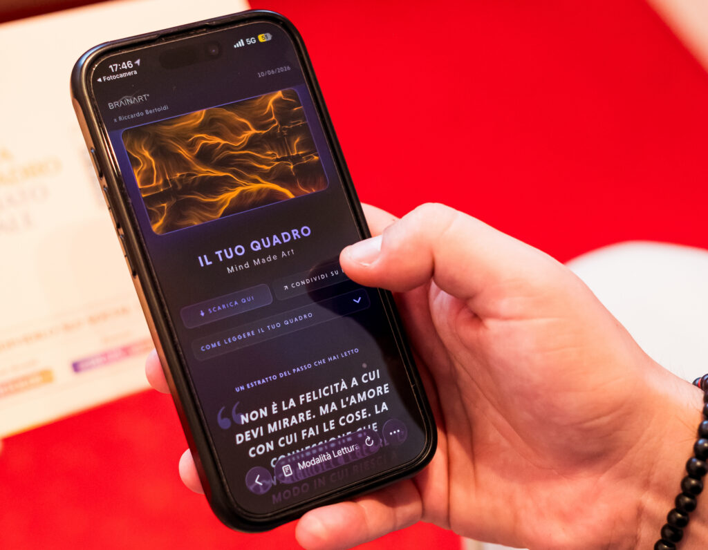 Una mano regge uno smartphone che mostra la pagina "IL TUO QUADRO – Mind Made Art" con il logo BrainArt® in alto: sotto un'opera a linee e vortici arancioni su fondo scuro, i pulsanti "Scarica qui", "Condividi su IG" e "Come leggere il tuo quadro", quindi un estratto del brano letto tra virgolette; in basso una barra con l'indicazione "Modalità Lettura". Sullo sfondo un piano rosso e un pieghevole chiaro appoggiato.
