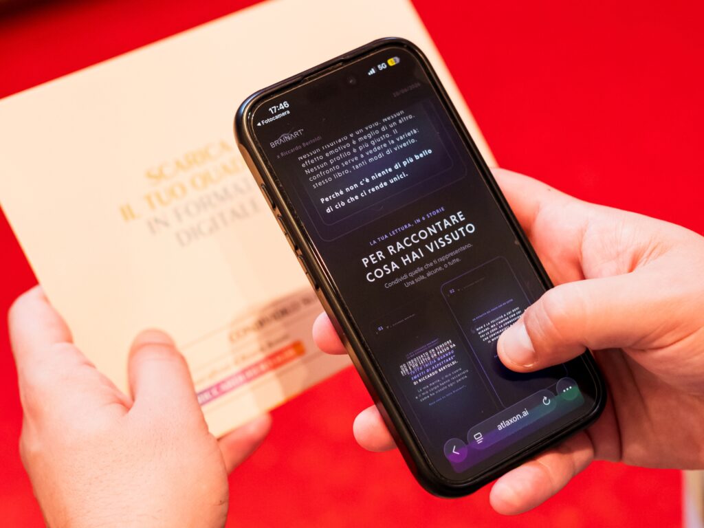Due mani reggono uno smartphone che mostra la sezione "Per raccontare cosa hai vissuto" della pagina BrainArt® di una sessione di lettura, con l'invito a condividere una o più storie e alcune anteprime di grafiche verticali con testo su fondo scuro; in alto un paragrafo ricorda che nessun risultato emotivo è migliore di un altro. Accanto, tenuto dall'altra persona, un pieghevole chiaro con la scritta "Scarica il tuo quadro in formato digitale". Sullo sfondo un piano rosso.