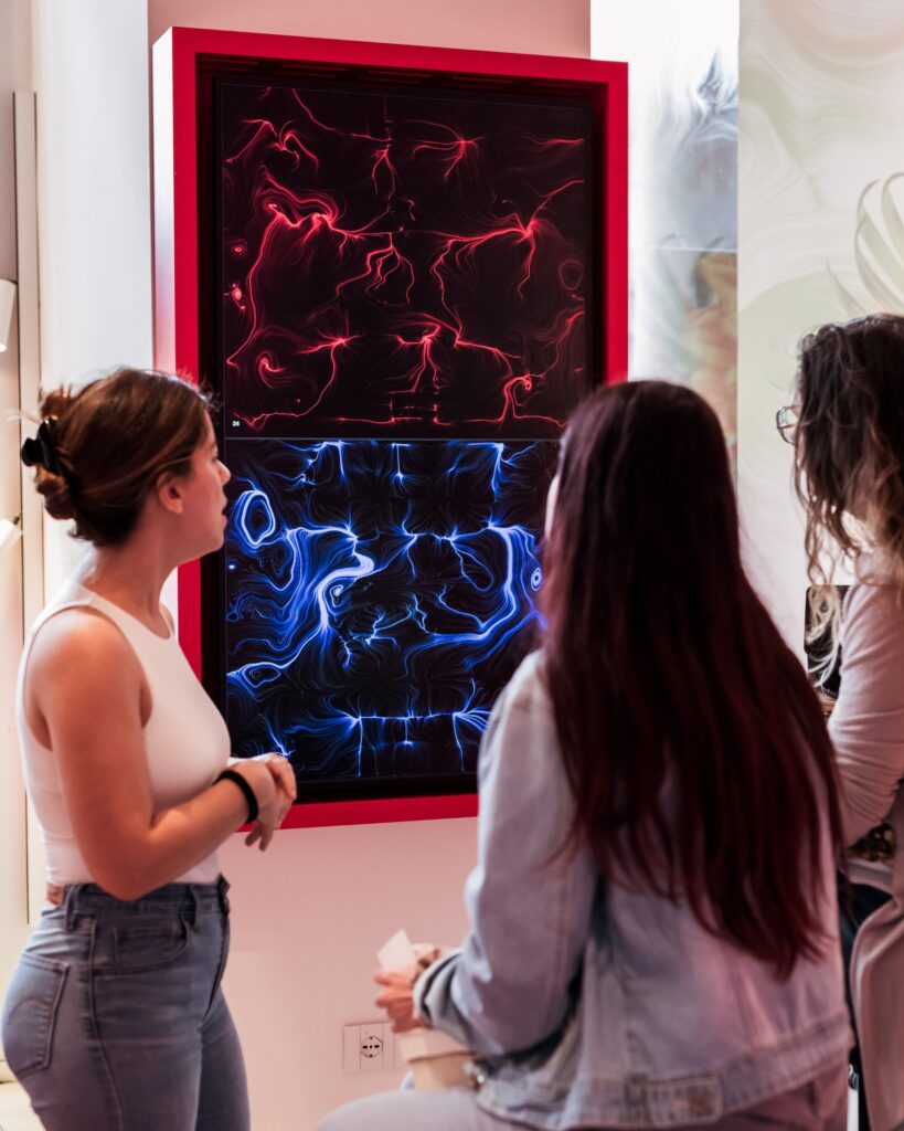 Un grande schermo verticale con cornice rossa, diviso in due parti, mostra due opere dalle linee e dai vortici, una rossa in alto e una blu in basso; tre donne lo osservano in un ambiente luminoso.