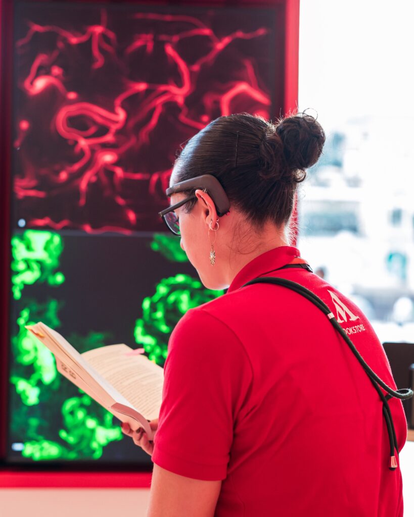Una donna di spalle, con una polo rossa che riporta il logo "M" dello store, occhiali e un sensore EEG sulla fronte, legge un libro aperto davanti a due schermi che mostrano opere dalle linee e dai vortici, uno rosso e uno verde.