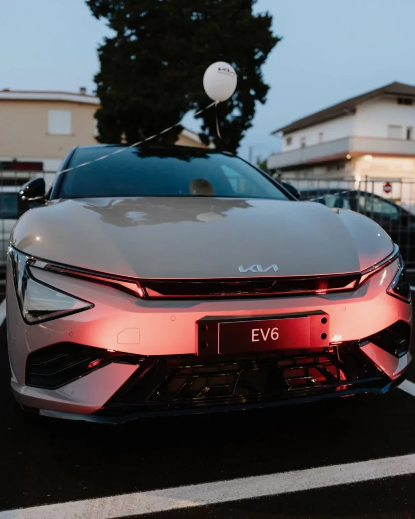 Una Kia EV6 grigia ripresa frontalmente al tramonto nel piazzale di una concessionaria, con i fari accesi che illuminano di rosso il muso e la scritta "EV6" al posto della targa; sopra il tetto è legato un palloncino bianco con il logo Kia, mentre sullo sfondo si vedono alcune abitazioni e un albero.
