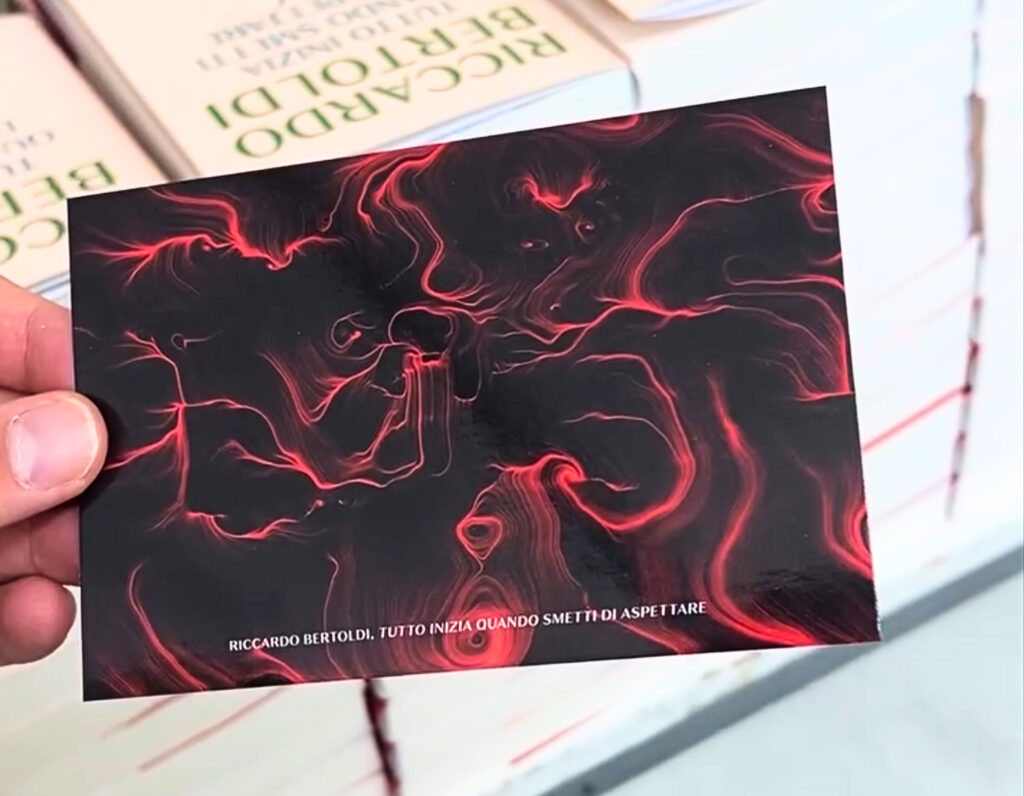 Una mano tiene una card con un'opera dalle linee e dai vortici rossi su fondo nero; in basso è stampato il nome Riccardo Bertoldi e il titolo del libro. Sullo sfondo, pile di copie del volume.