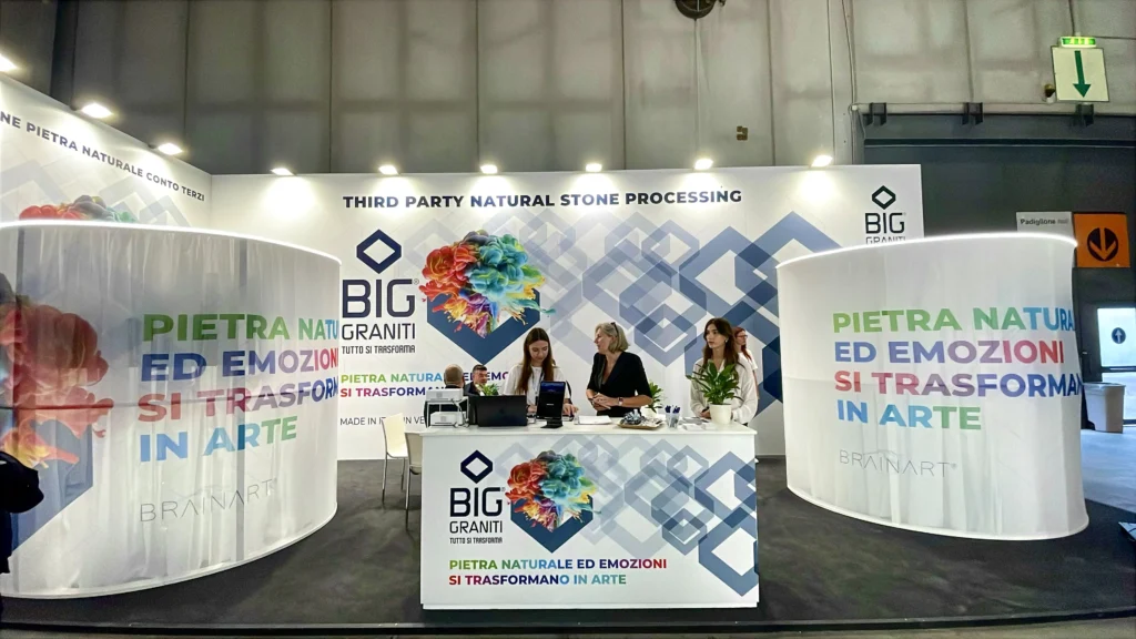 Stand fieristico con il logo BIG Graniti e il claim "Pietra naturale ed emozioni si trasformano in arte" ripetuto su due pannelli curvi bianchi; al bancone centrale, con il logo BrainArt, alcune persone in piedi; in alto la scritta "Third party natural stone processing".
