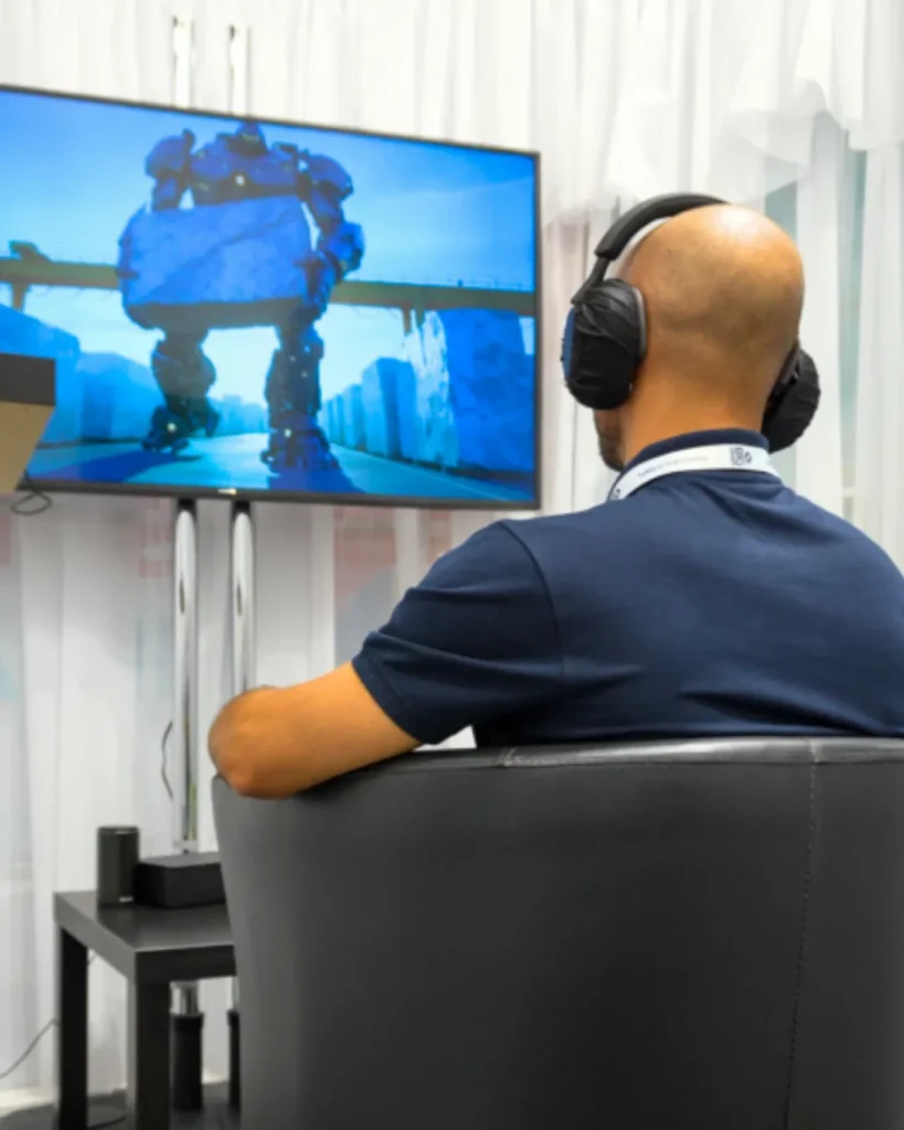 Un uomo di spalle, seduto in poltrona con cuffie e sensore EEG e pass al collo, guarda uno schermo appeso a parete che mostra un grande robot in stile animato su sfondo azzurro.
